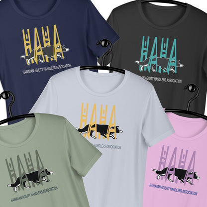Six short-sleeve graphic T-shirts on hangers, shown in navy, charcoal, light blue, sage green, and light pink. Each shirt features a large stylized 'HAHA' text in a different color above a black-and-white dog silhouette and the text HAWAIIAN AGILITY HANDLERS ASSOCIATION'.