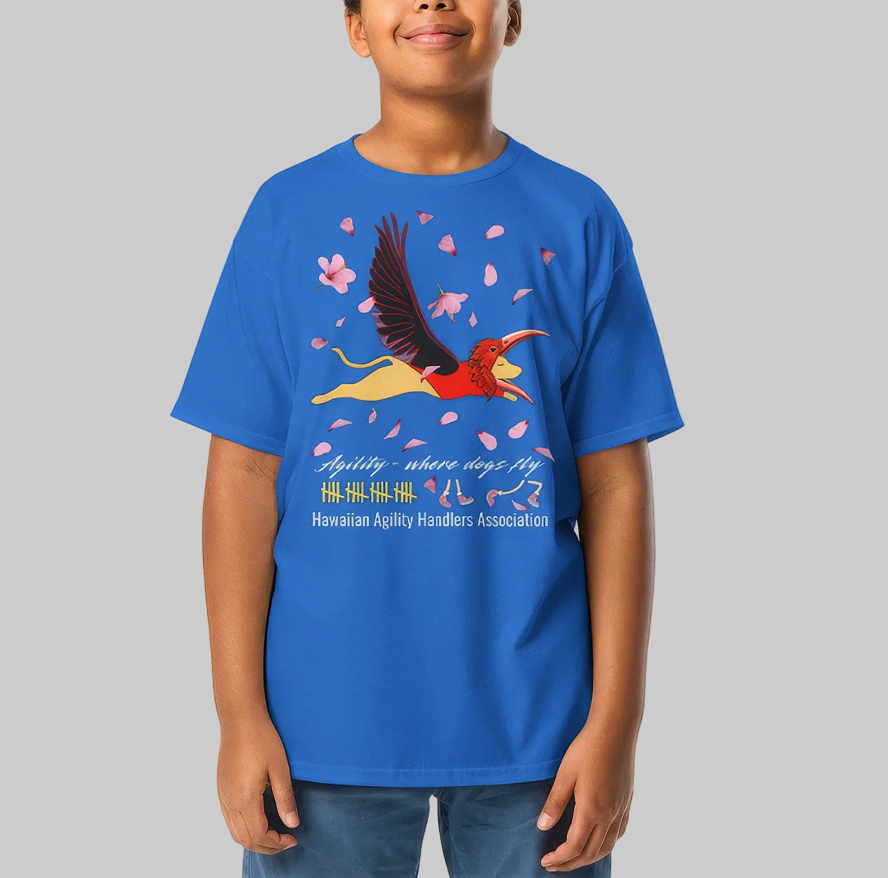 A child wearing a bright blue short-sleeve graphic T-shirt illustrating a dog wearing an I’iwi bird mask flying as pink hibiscus petals float through the air.  The text 'Agility - where dogs fly' and 'Hawaiian Agility Handlers Association' on the front, photographed against a plain gray background.
